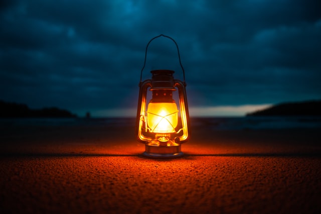 Imagem de um lampião ligado, durante a noite,  apoiado no chão com o céu e o mar de fundo.